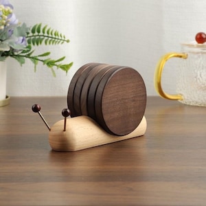 May include: A wooden snail-shaped coaster holder with six dark brown coasters. The snail's body is light-colored wood, and it has two antennae with red tips. The coasters are round and stacked.