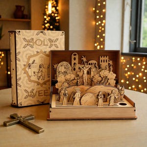 May include: A wooden book-shaped diorama depicting a religious scene with figures, trees, and architectural elements. The diorama is open, revealing the detailed laser-cut figures. A decorative book and a brass cross are also present.
