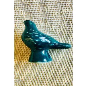 May include: A small, glossy, dark teal ceramic bird figurine standing on a flared pedestal base. The bird features a smooth, reflective surface with subtle, lighter teal mottled markings across its back and wings, captured in a side profile view.