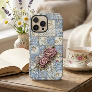 May include: A floral phone case with a patchwork design in shades of blue, white, and pink. The case features a cluster of pink flowers. The scene includes a teacup, a vase of flowers, and an open book on a wooden table.