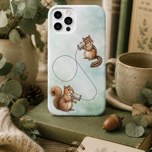 Könnte beinhalten: Eine weiße Handyhülle mit einem Aquarelldesign, das zwei Streifenhörnchen zeigt, die Blechdosen-Telefone benutzen. Der Hintergrund hat eine weiche grün-blaue Aquarellwaschung. Die Hülle ist von Büchern, Grünzeug und einer Tasse umgeben.