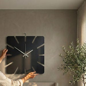 May include: A square, black wall clock with a minimalist design. The clock features a matte finish and cut-out hour markers. The hands are silver and the clock is being mounted on a neutral-colored wall. The overall aesthetic is modern and sleek.