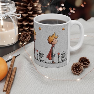 May include: White ceramic mug featuring a cartoon illustration of a character with yellow hair, a red cape, and a scarf. The design includes sheep, red flowers, and stars on a textured gray background. The mug has a white handle and is filled with a dark liquid.