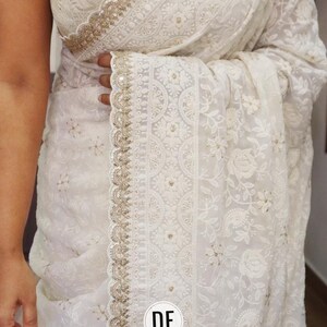 May include: A white saree featuring intricate floral embroidery and a scalloped border with gold sequin detailing. The fabric is sheer with a repeating pattern of small flowers. A circular white logo with the black letters DE is visible.