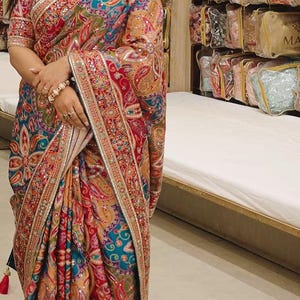 May include: A vibrant multi-coloured saree featuring intricate paisley patterns in shades of red, blue, gold, and green. The fabric is heavily embellished with metallic embroidery and small reflective mirror accents along the borders and body.