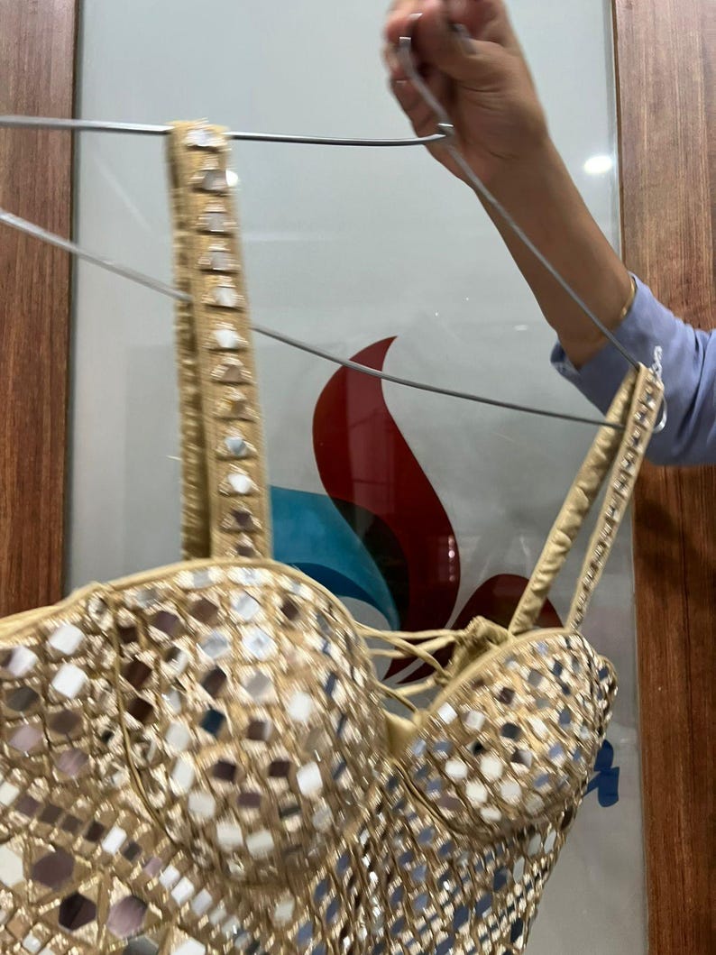 May include: A gold-colored bustier top with a mirrored design. The top has adjustable straps and a lace-up front. The bustier is hanging on a hanger.