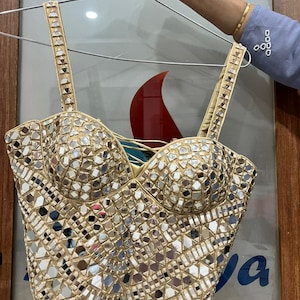 May include: A beige bustier top covered in small, reflective mirror pieces. The top has a sweetheart neckline and adjustable straps. The straps and edges are trimmed with small, decorative elements. The top is hanging on a metal hanger.