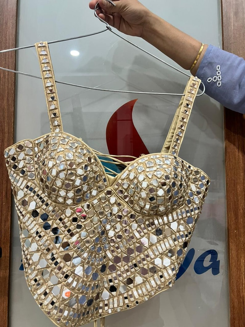 May include: A beige bustier top covered in small, reflective mirror pieces. The top has a sweetheart neckline and adjustable straps. The straps and edges are trimmed with small, decorative elements. The top is hanging on a metal hanger.