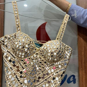 May include: A beige bustier top with a mosaic of small, reflective mirror pieces. The top has adjustable straps and a structured bodice. The garment is held up by a silver hanger.