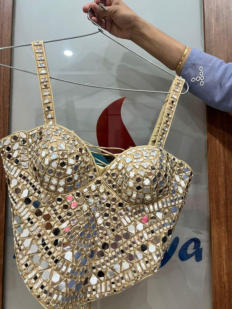 May include: A beige bustier top with a mosaic of small, reflective mirror pieces. The top has adjustable straps and a structured bodice. The garment is held up by a silver hanger.