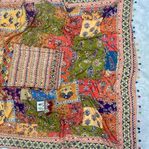 May include: A colorful patchwork textile with a variety of patterns and colors, including orange, green, yellow, and blue. The textile features floral and paisley designs, with a decorative border and small mirrored accents. The edges are trimmed with small, colorful pom-poms.