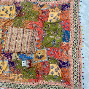 May include: A colorful patchwork textile with a variety of patterns and colors, including orange, green, yellow, and blue. The textile has a decorative border with small mirrors and tassels. The design features floral and paisley motifs.