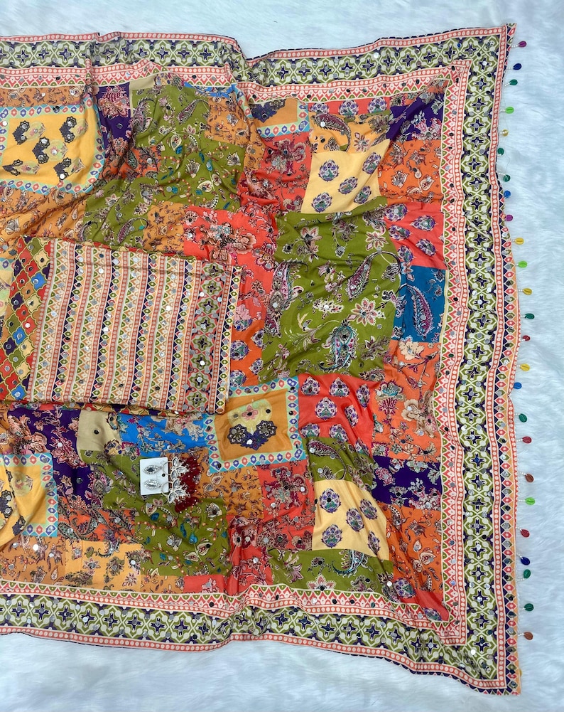 May include: A colorful patchwork textile with a variety of patterns and colors, including orange, green, yellow, and blue. The textile has a decorative border with small mirrors and tassels. The design features floral and paisley motifs.