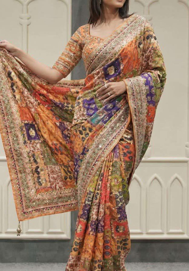 May include: A vibrant, patterned saree in shades of orange, purple, and green. The fabric features intricate designs and is embellished with shimmering details along the borders. The saree's design includes a matching blouse with similar patterns.