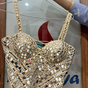 May include: A beige bustier top covered in small, reflective mirror pieces. The top has a sweetheart neckline and adjustable straps. The straps and edges are trimmed with small, decorative elements. The top is hanging on a metal hanger.