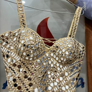 May include: A gold-colored bustier top with intricate mirror embellishments. The top features adjustable straps and a sweetheart neckline. The surface is covered in small, reflective mirror pieces, creating a shimmering effect. The garment is held up by a silver hanger.