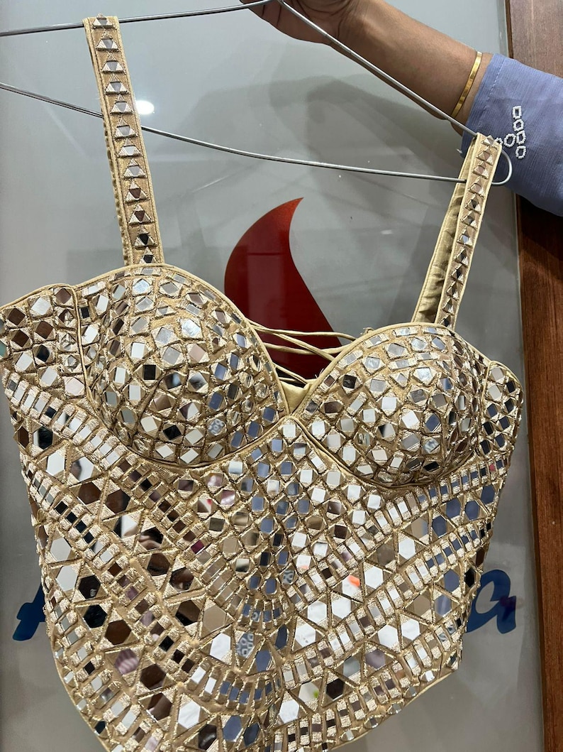 May include: A gold-colored bustier top with intricate mirror embellishments. The top features adjustable straps and a sweetheart neckline. The surface is covered in small, reflective mirror pieces, creating a shimmering effect. The garment is held up by a silver hanger.