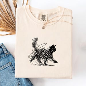 Puede incluir: Camiseta color crema con un diseño de gato negro estilo garabato. El gato se representa caminando, con cola y cabeza. La camiseta está doblada y tiene una etiqueta Comfort Colors.