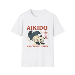 Puede incluir: Camiseta blanca con un panda de dibujos animados practicando Aikido. El panda lleva un gi azul y negro. El texto "AIKIDO" es rojo, con caracteres japoneses y "PRACTICING UKEMI" debajo.
