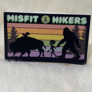 May include: A rectangular sticker featuring the words "MISFIT HIKERS" in green bubble letters. The sticker depicts silhouettes of a bison, an alien, and Bigfoot walking together against a sunset backdrop with trees.