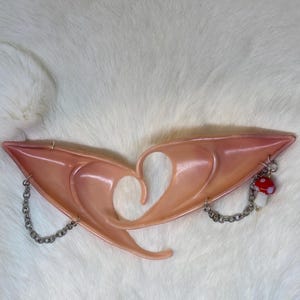 May include: A pair of pink elf ears with a heart-shaped cutout in the center. The ears are connected by a silver chain and adorned with a small mushroom charm. The ears are laying on a white, fluffy surface.