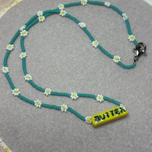 May include: A beaded necklace featuring a teal chain adorned with white and yellow daisy-like flowers. A yellow rectangular pendant with the word "BUTTER" in blue lettering is at the bottom of the necklace. The necklace has a silver clasp.