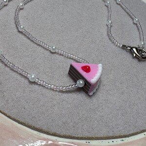 May include: A delicate necklace featuring a slice of cake pendant. The necklace has a beaded chain with small white pearls and a lobster clasp. The cake pendant is pink with white frosting and a red cherry on top.