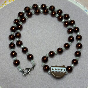 May include: A two-strand necklace featuring alternating dark brown and white faux pearls. The necklace has a brown pendant with a white and blue design. A silver clasp secures the necklace.