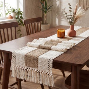 Può includere: Runner da tavolo rettangolare a righe crema e marroni con frange. Il runner è realizzato in tessuto intrecciato ed è posto su un tavolo di legno marrone scuro. Una candela e un vaso con fiori secchi sono sul tavolo.