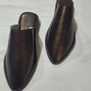 Könnte beinhalten: Ein Paar bronzefarbene Leder-Schlupfschuhe. Die Schuhe haben eine spitze Zehenpartie und einen niedrigen Absatz. Die Kanten sind mit einem goldfarbenen Material verziert. Die Schuhe werden auf einer weißen Oberfläche präsentiert.