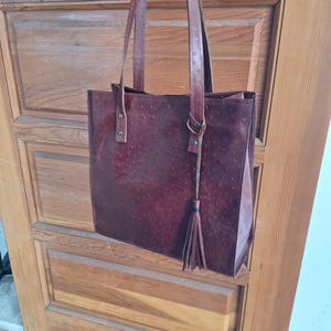 May include: A burgundy leather tote bag with a textured finish and long shoulder straps. The bag features a rectangular shape, a tassel detail, and is hanging against a wooden door.