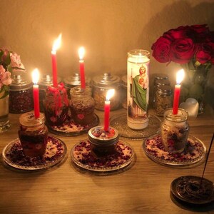 以下が含まれることがあります： ガラス瓶に入った赤いろうそくが灯り、ガラス皿にバラの花びらが散りばめられたスピリチュアルなアレンジメント。聖人のイメージが描かれた大きなろうそく、赤いバラの花束、ハーブの入った瓶が並んでいます。