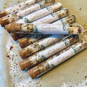 Op de afbeelding: Een verzameling glazen buisjes gevuld met badzout en botanische ingrediënten. Elk buisje heeft een kurk en een label met het woord "Schön" en de zin "Dass Du Da Bist". De zouten zijn gemengd met kruiden en bloemblaadjes.