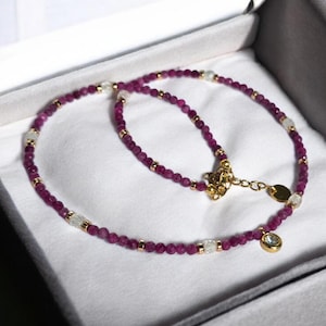 May include: A necklace with small, round, deep pink beads, clear faceted beads, and gold-coloured accents. The necklace is displayed in a white box, highlighting its elegant design. A small, gold-coloured charm with a clear stone hangs from the clasp.