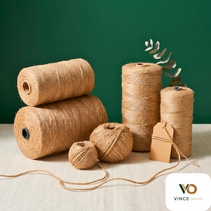 May include: A selection of natural twine spools and balls in different sizes. The spools are cylindrical, and the balls are round. A blank brown tag hangs from one of the spools. The twine is a light brown colour, and the background is a dark green.