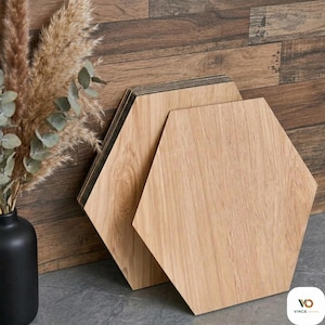 May include: Two light-colored, hexagonal wooden tiles are displayed, showcasing a natural wood grain pattern. The tiles are leaning against a wooden wall, with a black vase holding dried plants in the background. The tiles are a light brown color.