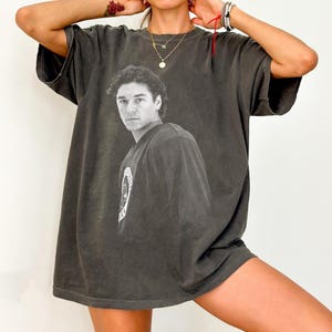 May include: Dark grey oversized t-shirt with a black and white portrait of a man. The shirt has a distressed appearance and a crew neck. A circular graphic is visible on the left sleeve. A casual, unisex garment.