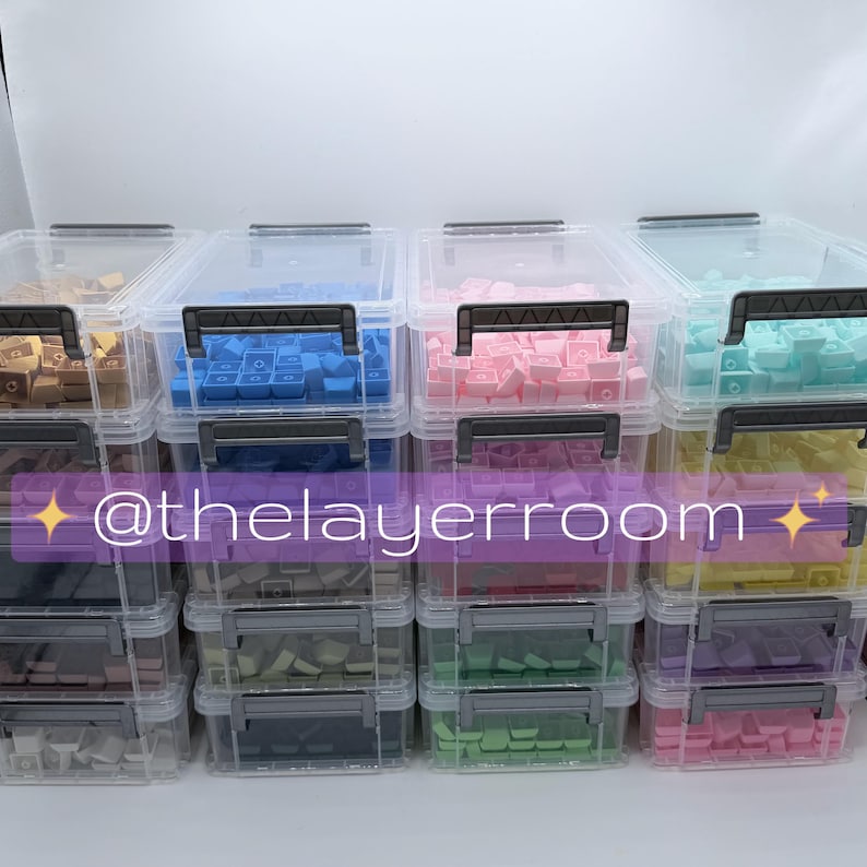Peut inclure: Plusieurs bo&icirc;tes de rangement en plastique transparent remplies de touches de clavier color&eacute;es. Les touches sont de diff&eacute;rentes couleurs, dont le bleu, le rose et le vert. Chaque bo&icirc;te a une poign&eacute;e noire et est empil&eacute;e. Le texte "@thelayerroom" est visible.