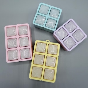 May include: Four silicone ice cube trays in pastel colors including pink, light blue, lavender, and yellow. The pink and yellow trays have six square compartments, while the blue and lavender trays have four square compartments, all filled with ice.