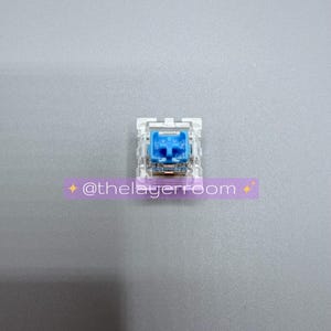 Peut inclure: Un interrupteur de clavier m&eacute;canique unique avec un bo&icirc;tier transparent et une tige bleu vif. L'interrupteur est centr&eacute; sur une surface grise. Un autocollant violet avec le texte "@thelayerroom" est dans l'image.