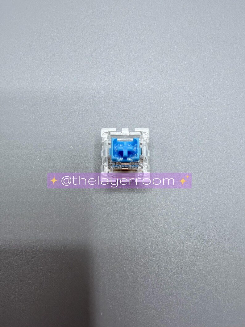 Peut inclure: Un interrupteur de clavier m&eacute;canique unique avec un bo&icirc;tier transparent et une tige bleu vif. L'interrupteur est centr&eacute; sur une surface grise. Un autocollant violet avec le texte "@thelayerroom" est dans l'image.