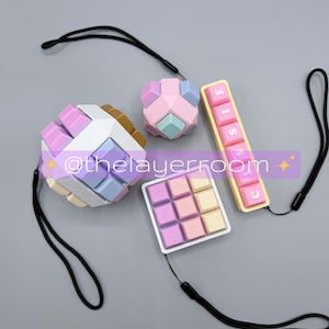 May include: Four colorful puzzle toys with black lanyards. The puzzles include a cube, a smaller cube, a square grid, and a rectangular piece with the word "CLASSIC". The toys are in pastel colors of pink, blue, yellow, and white.