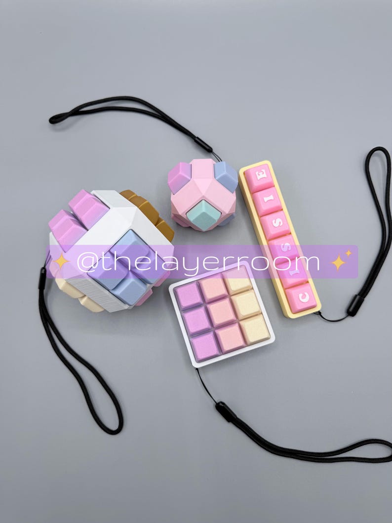 Peut inclure: Quatre jouets de puzzle color&eacute;s avec des lani&egrave;res noires. Les puzzles comprennent un cube, un plus petit cube, une grille carr&eacute;e et une pi&egrave;ce rectangulaire avec le mot "CLASSIC". Les jouets sont dans des couleurs pastel rose, bleu, jaune et blanc.