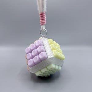 May include: A cube-shaped charm featuring a grid of pastel-colored plastic squares in lavender, pale yellow, and mint green. The cube is framed by a silver-toned border and hangs from a white cord with a pink wrapped accent and a metal ring.