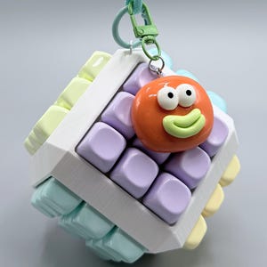 Peut inclure: Un jouet anti-stress en forme de cube avec un cadre blanc incrusté de touches de clavier aux couleurs pastel, notamment lavande, vert menthe et jaune pâle. Une breloque orange en forme de visage souriant est fixée à un clip métallique vert clair.