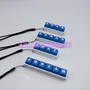 Peut inclure: Quatre porte-cl&eacute;s personnalis&eacute;s en forme de touches de clavier, avec les noms "AMAR", "ETHAN", "AT" et "JOSIAH". Chaque porte-cl&eacute;s a des touches bleues avec des lettres blanches, mont&eacute;es sur une base blanche et attach&eacute;es &agrave; un cordon noir. Le fond est blanc.