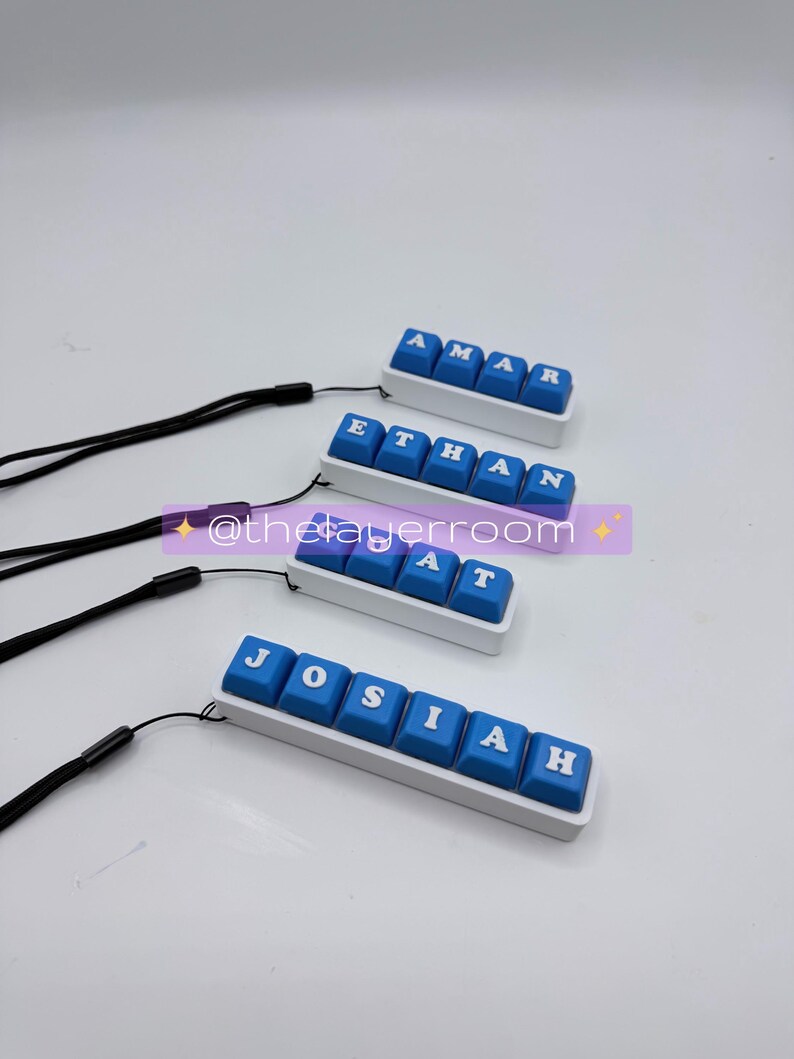 Peut inclure: Quatre porte-cl&eacute;s personnalis&eacute;s en forme de touches de clavier, avec les noms "AMAR", "ETHAN", "AT" et "JOSIAH". Chaque porte-cl&eacute;s a des touches bleues avec des lettres blanches, mont&eacute;es sur une base blanche et attach&eacute;es &agrave; un cordon noir. Le fond est blanc.