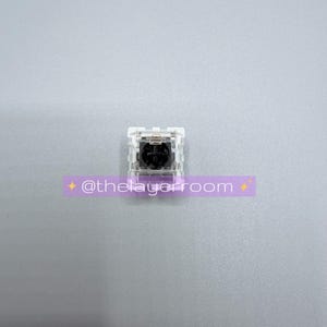 Peut inclure: Un interrupteur de clavier m&eacute;canique dot&eacute; d'un bo&icirc;tier transparent et d'une tige noire avec une fixation en croix. Une superposition rectangulaire violette est plac&eacute;e sous l'interrupteur, affichant le texte blanc @thelayerroom.