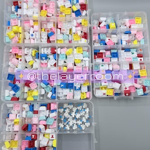 Peut inclure: Assortiment de touches de clavier color&eacute;es dans des organisateurs en plastique transparent. Les touches sont de diff&eacute;rentes couleurs, notamment rose, bleu, jaune et blanc. Certaines touches comportent des lettres et des symboles. Le texte "@thelayerroom" est inclus.