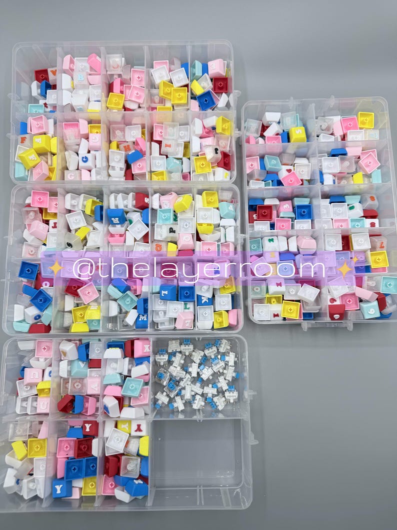 Peut inclure: Assortiment de touches de clavier color&eacute;es dans des organisateurs en plastique transparent. Les touches sont de diff&eacute;rentes couleurs, notamment rose, bleu, jaune et blanc. Certaines touches comportent des lettres et des symboles. Le texte "@thelayerroom" est inclus.